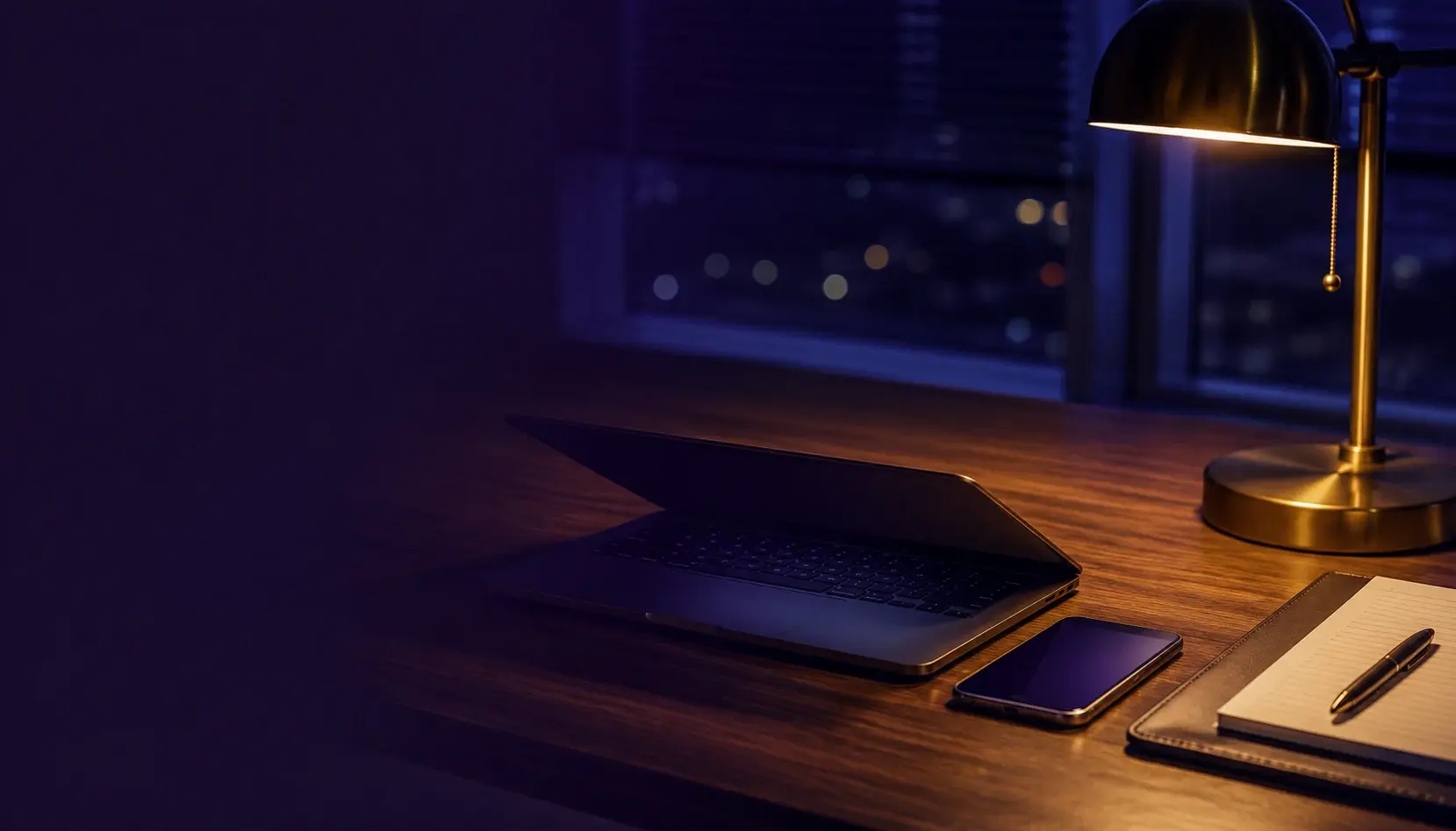 An office desk at night with a laptop lid half closed and a phone in hand, one lamp on