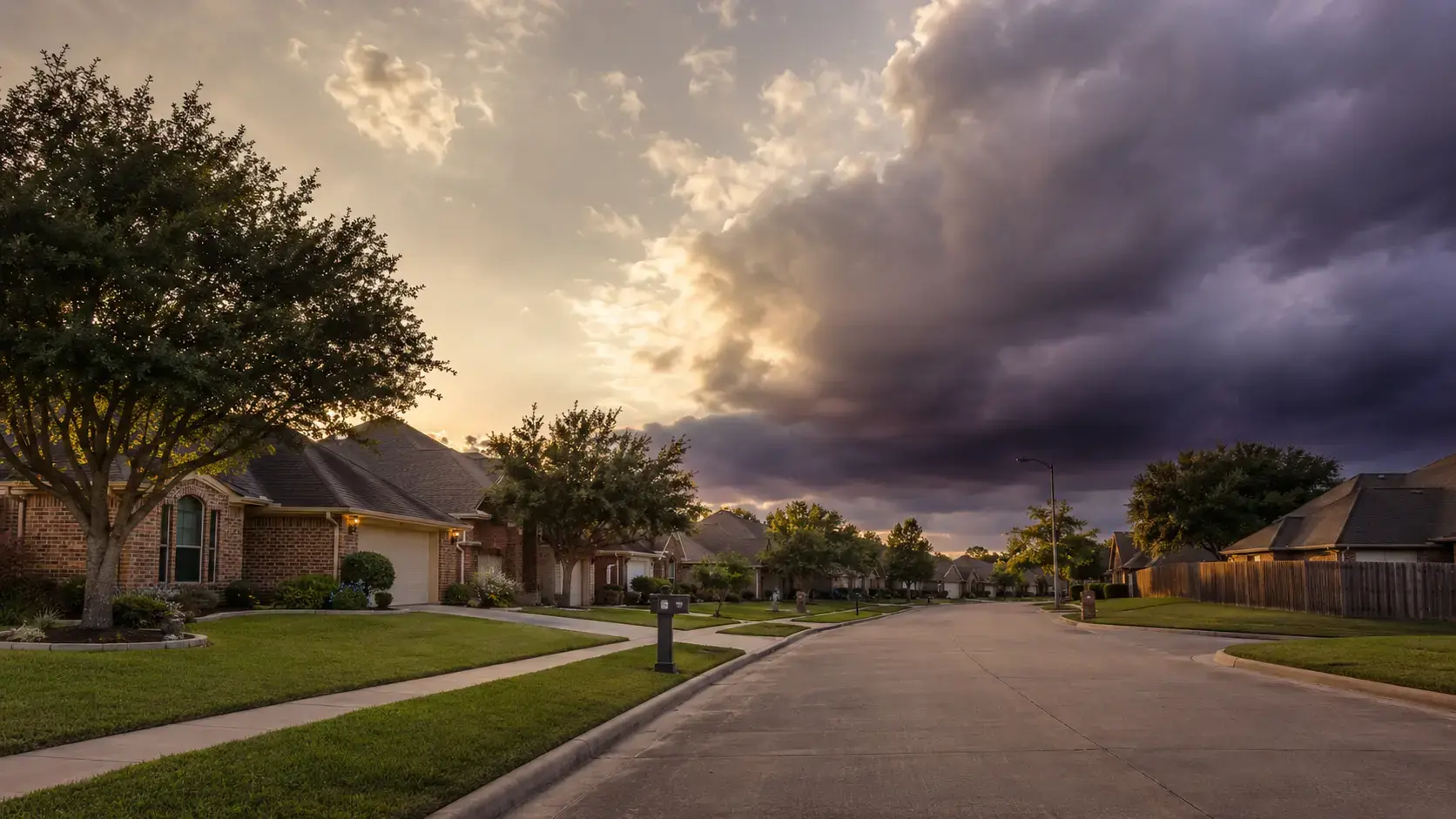 A quiet Houston residential street of brick homes and live oaks in golden evening light as storm clouds build on the horizon